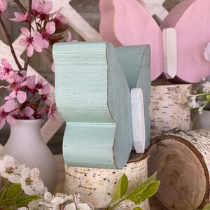 Pu&ograve; includere: Due decorazioni in legno a forma di farfalla, una dipinta di blu chiaro e l'altra di rosa, sono esposte su fette di legno di betulla. Le farfalle sono posizionate con le ali aperte, creando un'esposizione fantasiosa e decorativa.
