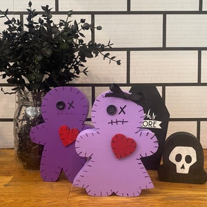 May include: Two wooden voodoo doll figures in purple and lavender, each with a red heart, stitched details, and button eyes. A black coffin-shaped sign and a skull-shaped figure are also present, with a black plant in a glass jar in the background. The word "more" is visible.