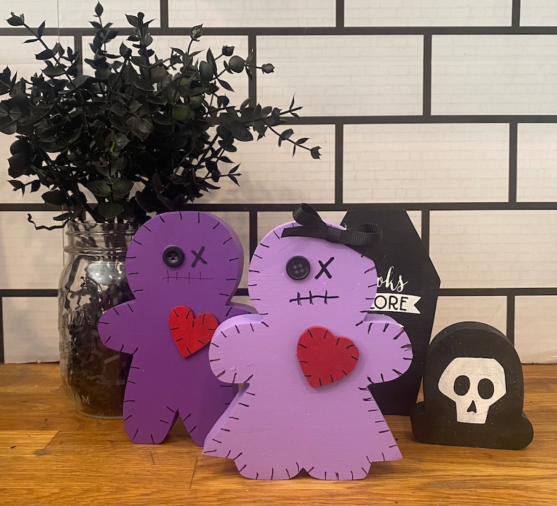 May include: Two wooden voodoo doll figures in purple and lavender, each with a red heart, stitched details, and button eyes. A black coffin-shaped sign and a skull-shaped figure are also present, with a black plant in a glass jar in the background. The word "more" is visible.