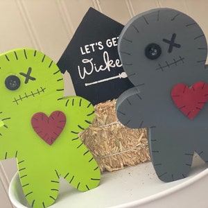 May include: Two wooden voodoo doll figures, one lime green and one gray, with stitched details, button eyes, and red heart accents. A small hay bale and a sign that says "LET'S GET Wicked" are in the background. Halloween decor.