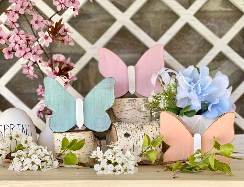 Pu&ograve; includere: Tre figurine di farfalla in legno, dipinte in rosa pastello, azzurro chiaro e pesca, sono esposte su fette di legno di betulla. Le farfalle sono disposte in fila, con la farfalla rosa a sinistra, la farfalla azzurra al centro e la farfalla pesca a destra. Le farfalle sono tutte rivolte nella stessa direzione, con le ali spiegate.
