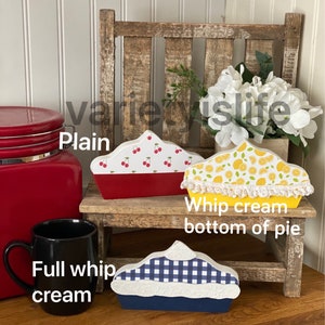 May include: Three wooden pie-shaped decorations, each with a different design. The first pie is red with white cherries, the second is yellow with lemons and a white lace trim, and the third is blue with a white and blue checkered pattern. The pies are sitting on a wooden stool.