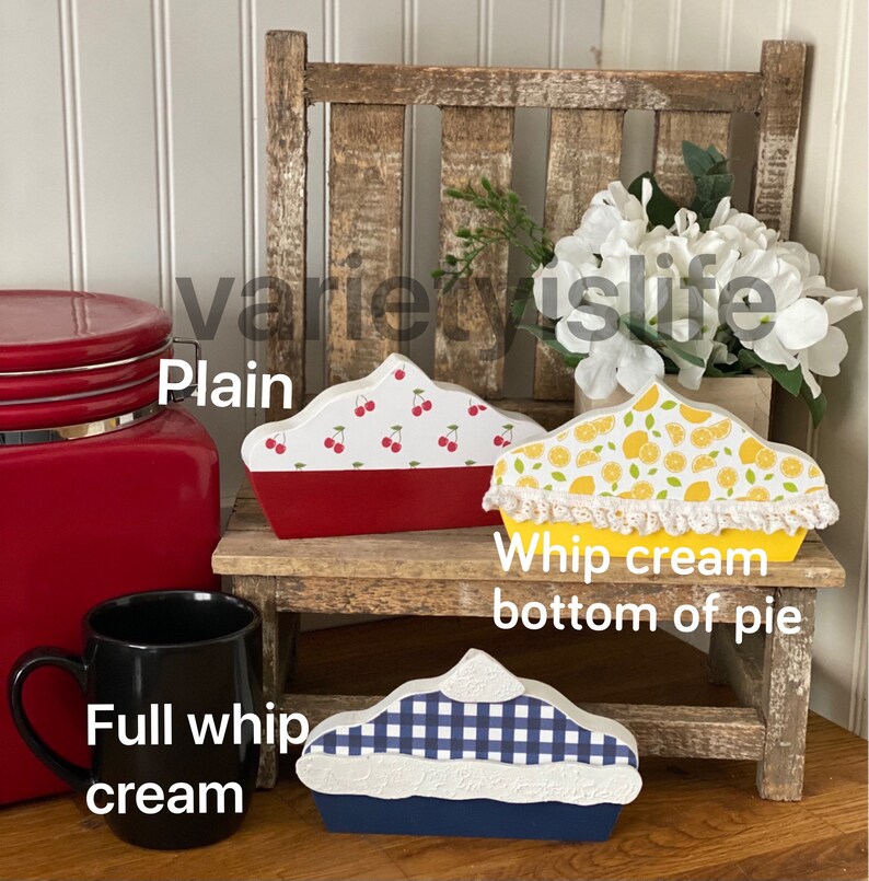May include: Three wooden pie-shaped decorations, each with a different design. The first pie is red with white cherries, the second is yellow with lemons and a white lace trim, and the third is blue with a white and blue checkered pattern. The pies are sitting on a wooden stool.