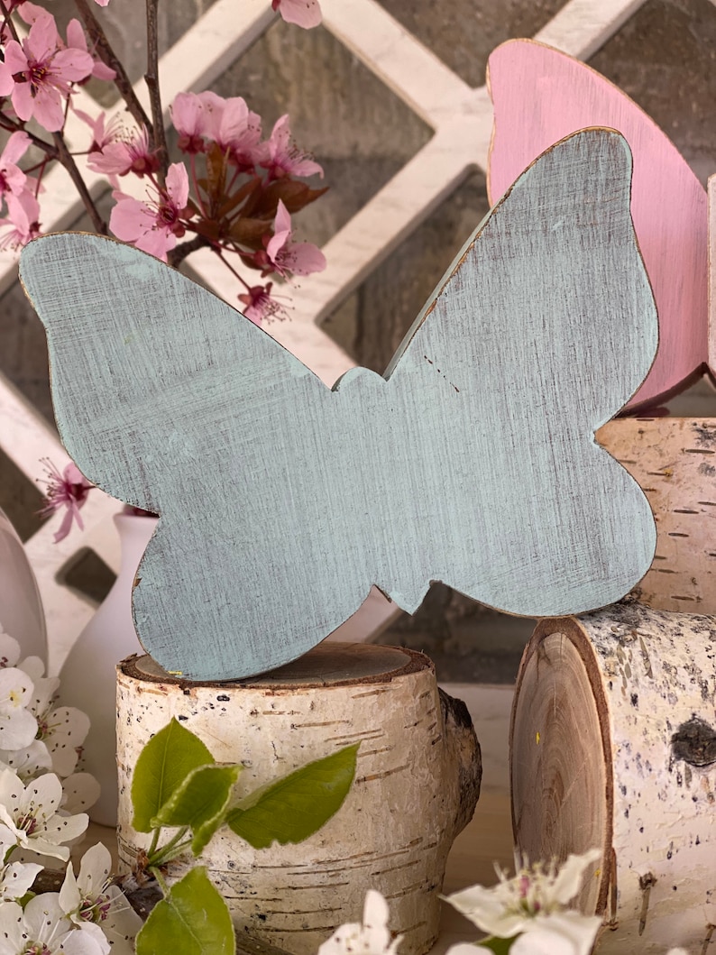 Pu&ograve; includere: Un'immagine di una farfalla di legno blu chiaro con una finitura usurata, seduta su un tronco di betulla. La farfalla &egrave; su una superficie bianca con fiori rosa.
