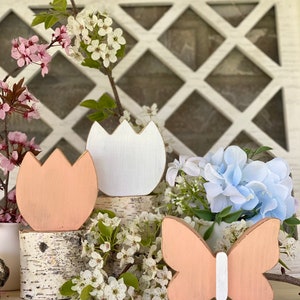 Pu&ograve; includere: Tre decorazioni in legno a tema primaverile: un tulipano color pesca, un tulipano bianco e una farfalla color pesca con corpo bianco. Le decorazioni sono disposte su un tronco di betulla e circondate da fiori bianchi e blu.