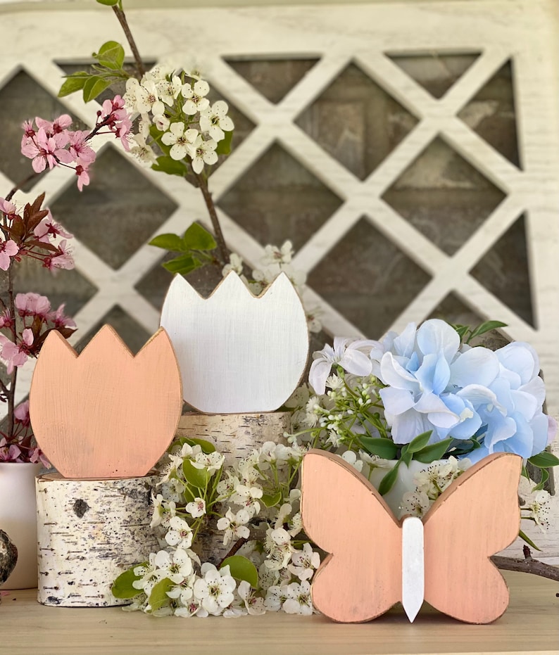 Pu&ograve; includere: Tre decorazioni in legno a tema primaverile: un tulipano color pesca, un tulipano bianco e una farfalla color pesca con corpo bianco. Le decorazioni sono disposte su un tronco di betulla e circondate da fiori bianchi e blu.