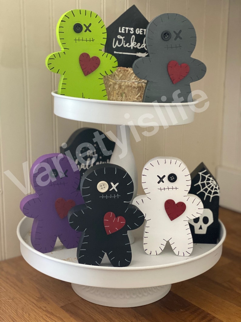 May include: A two-tiered white tray displays Halloween-themed wooden figures. The figures include voodoo doll shapes in green, gray, purple, black, and white, each with a red heart and stitched details. Decorative signs and a small bale of hay complete the arrangement.