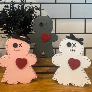 May include: Three wooden gingerbread-style figures in pink, gray, and white. Each has a stitched outline, button eyes, and a stitched mouth. A red heart is affixed to the chest, and a black bow is on the pink and white figures.