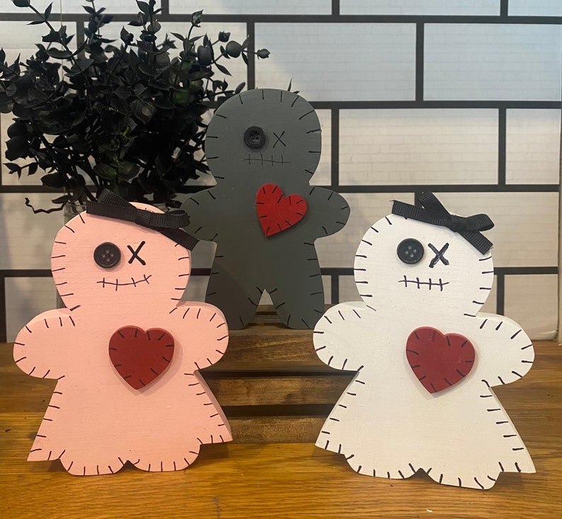 May include: Three wooden gingerbread-style figures in pink, gray, and white. Each has a stitched outline, button eyes, and a stitched mouth. A red heart is affixed to the chest, and a black bow is on the pink and white figures.