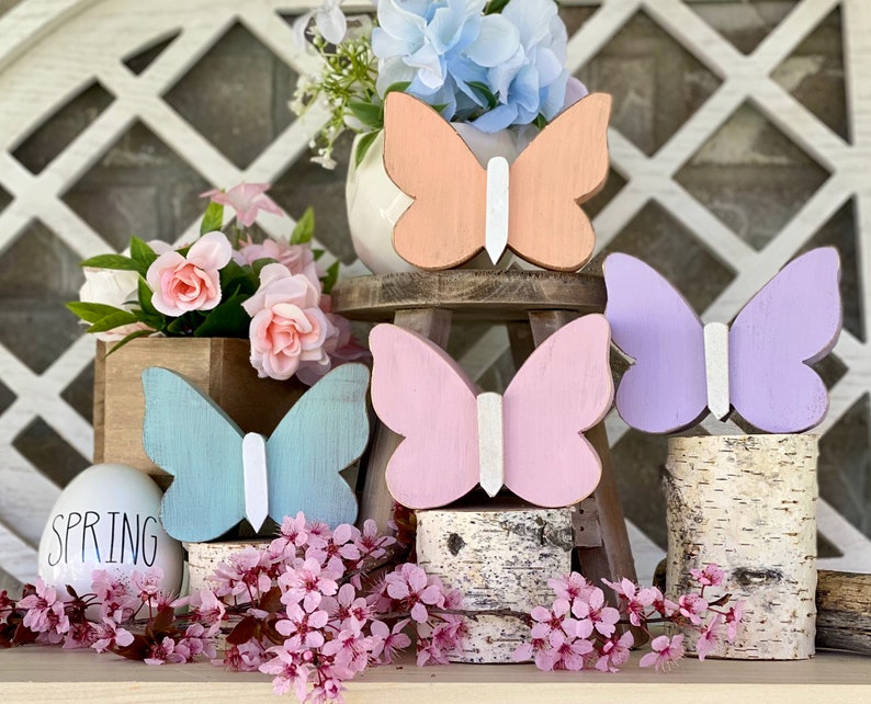 Pu&ograve; includere: Quattro decorazioni a forma di farfalla in legno in colori pastello, rosa, viola, blu e pesca, con accenti bianchi. Le farfalle sono esposte su un supporto a pi&ugrave; livelli con basi in corteccia di betulla. La parola "SPRING" &egrave; scritta su un uovo bianco.