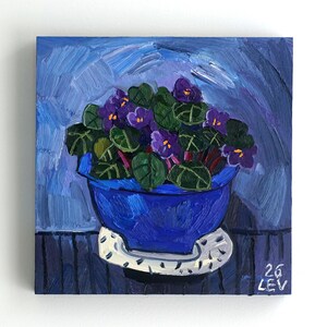 Könnte beinhalten: Original Ölgemälde einer Topfpflanze mit afrikanischen Veilchen in einem blauen Topf auf einer weißen Untertasse. Das Gemälde zeigt violette Blüten und grüne Blätter vor blauem Hintergrund. Die Signatur des Künstlers, "26 LEV", ist unten rechts zu sehen.
