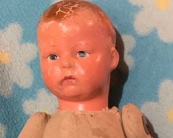 May include: A vintage porcelain doll with a cracked face, wearing a beige jumpsuit and black boots. The doll has blue eyes and is posed with one hand on its hip.