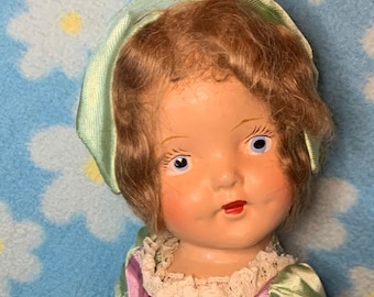 May include: A vintage doll with brown hair, wearing a light green and purple dress with a green bow. The doll is wearing white socks and black shoes.