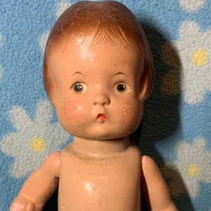 May include: A vintage porcelain doll with a painted face, brown hair, and a nude body. The doll has a cracked surface and is missing paint in some areas.