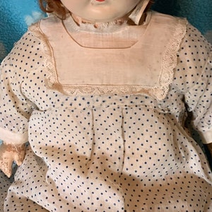 May include: A vintage porcelain doll wearing a white dress with blue polka dots and a white lace trim. The doll has red hair and a painted face.