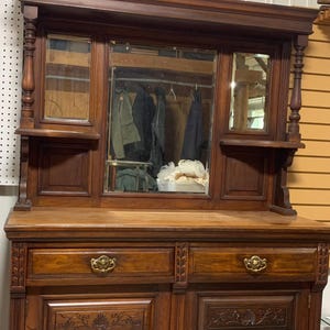 May include: A dark brown antique wooden buffet with a mirror. The buffet has two drawers with ornate brass handles and two cabinet doors with carved details. The mirror is flanked by small shelves and decorative columns. The buffet is a classic piece of furniture.