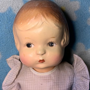 May include: A vintage porcelain doll with painted brown hair, closed eyes, and a painted pink blush on her cheeks. She is wearing a light purple dress with white trim.