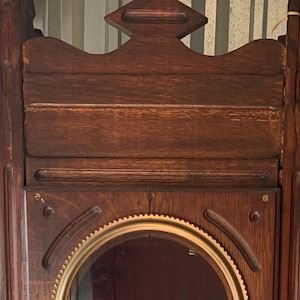 May include: Antique wooden grandfather clock with a dark brown finish. Features a round clock face with a gold-colored trim, a small oval opening, and a lower cabinet door with a brass knob. A classic piece of furniture.
