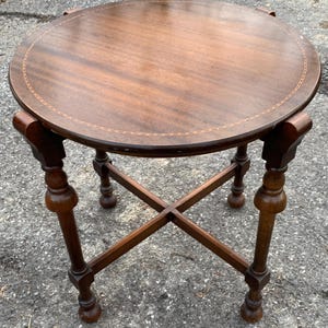 May include: A round, antique wooden side table with a dark brown finish. The table has a decorative inlaid border around the top and four ornate legs with a cross-shaped base. The table is approximately 24 inches in diameter.