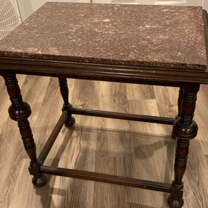 May include: A dark brown, antique side table with a rectangular top. The tabletop is a speckled, reddish-brown stone. The table has ornate, turned legs and a lower shelf for added stability. A classic piece of furniture.