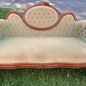 May include: An antique, light green velvet sofa with a wooden frame. The sofa features a tufted back and rolled arms. The wooden frame is a dark brown color. The sofa is placed on a grassy surface.