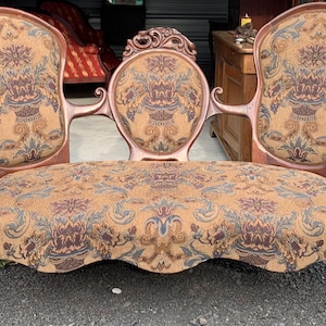 May include: A wooden settee featuring a carved frame and three distinct backrests upholstered in a tan fabric with a multi-colored floral and scroll pattern. The seat is continuous and upholstered in the same patterned fabric, supported by curved legs.