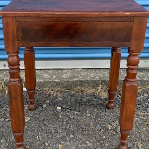 May include: A dark brown wooden table with a single drawer and turned legs. The table has a simple, rustic design.