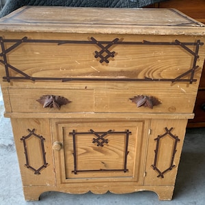 May include: A wooden cabinet with a light brown finish and dark brown painted details. The cabinet has a single drawer and a door with a single shelf. The door and drawer have a geometric design painted on them.