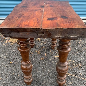 May include: A rustic wooden table with a rectangular top and four turned legs. The table top is made of two pieces of wood that have been joined together. The table is made of dark brown wood and has a distressed finish.