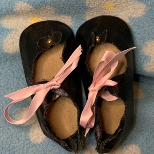 May include: A pair of black leather baby shoes with pink satin ribbons tied around the ankles. The shoes have a gold button on each.