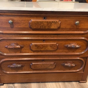 May include: A wooden chest of drawers featuring a white marble top and three horizontal drawers. Each drawer is accented with a central rectangular panel, a keyhole, and decorative metal hardware. The wood has a warm brown finish.
