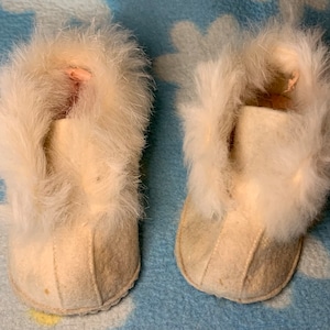 May include: A pair of white baby booties with fluffy white fur trim. The booties are made of a soft, white fabric.