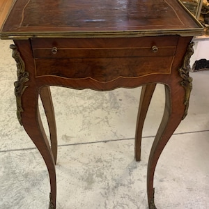 May include: A small, antique wooden side table with a rectangular top and a single drawer. The table features curved legs and ornate brass accents. The wood has a rich, dark brown finish with a decorative inlay.