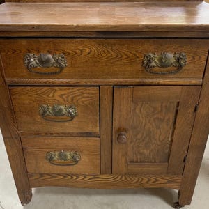 May include: A vintage wooden cabinet with a light brown finish. It features two drawers with ornate handles, two smaller drawers, and a cabinet door with a round knob. The cabinet sits on small wheels.