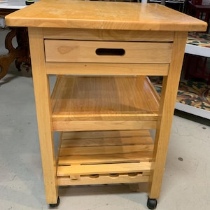 May include: A light-colored wooden kitchen cart with a drawer, two shelves, and a slatted bottom shelf for storage. The cart has a rectangular top and is on wheels for easy mobility. The cart is made of light-colored wood.