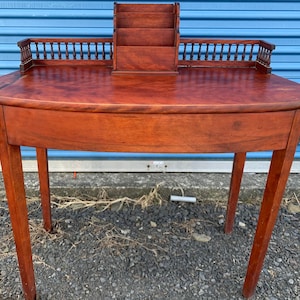 May include: A mahogany wood writing desk with a curved front and a small letter holder on top. The desk has a simple design with turned legs and a decorative railing.