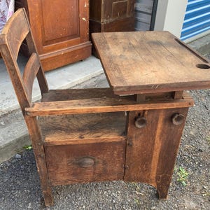 May include: A vintage wooden school desk with an attached chair, featuring a rectangular writing surface with a circular inkwell hole and a groove for pens. The base includes a storage compartment with a drawer and two round wooden knobs.