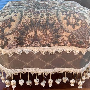May include: An upholstered ottoman with a patterned top in shades of beige, brown, and green. The base is a darker brown with a diamond pattern, trimmed with cream-colored fringe and tassels. The ottoman sits on wooden legs.