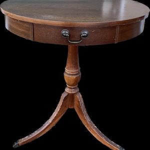 May include: A vintage, oval-shaped wooden side table with a single drawer and a dark finish. The table has a pedestal base with three curved legs. The drawer features a metal handle. The table is approximately 28 inches in diameter.