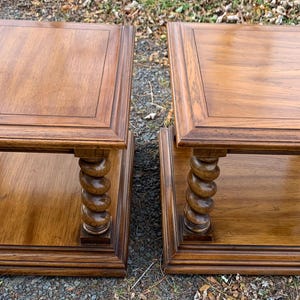 May include: Two square wooden side tables with a warm brown finish. Each table features a lower shelf and decorative, turned legs. The tops have a framed edge. A classic furniture set.