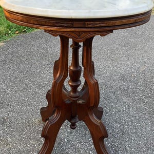 May include: A round side table featuring a white marble top with grey veining. The base is constructed from dark brown wood, showcasing an intricate pedestal design with curved legs and a central turned column support beneath the tabletop.