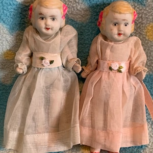 May include: Two porcelain dolls wearing white dresses with pink sashes and pink bows in their hair. The doll on the left is wearing a light blue dress and the doll on the right is wearing a pink dress.