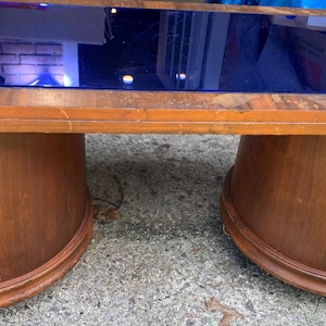 May include: A rectangular coffee table featuring a medium-brown wooden frame and a smooth, dark blue glass top. The table is supported by two curved, semi-circular wooden pedestals, each equipped with small wheels at the base for mobility.
