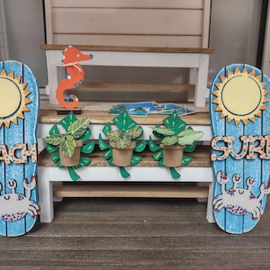 Puede incluir: Un banco de madera con tres plantas en macetas y dos letreros de chanclas azules y blancas. Un letrero dice "Beach" y el otro dice "Surf".