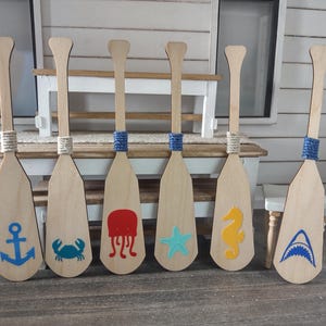 May include: Six wooden miniature oars with nautical-themed designs. Each oar features a different sea creature or symbol, including an anchor, crab, jellyfish, starfish, seahorse, and shark. The handles are wrapped with twine or blue rope.