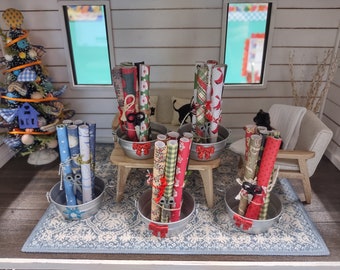 May include: Five miniature metal buckets filled with rolls of colourful wrapping paper, each bucket has a pair of scissors and a red bow. The buckets are on a blue and white patterned rug in a dollhouse setting.