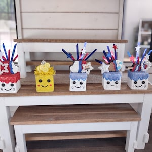 May include: A collection of miniature decorative boxes with smiling faces. The boxes are painted red, yellow, and blue, and topped with various embellishments like stars, ribbons, and pearls. The boxes are arranged on a small wooden table.