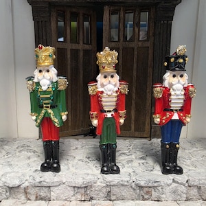 May include: Three nutcracker figurines standing in a row. The left figure wears a green coat and gold crown. The centre figure wears a red coat and gold crown. The right figure wears a red coat and black crown. All have white beards and black boots.