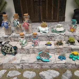 May include: A collection of miniature turtle-themed decorative items including glass bottles filled with sand, beads, or stones, and small turtle figurines, some featuring air plants or the word SEA, arranged on a stone-textured tiered display.