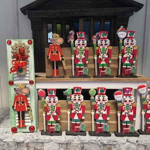 May include: A collection of wooden nutcracker and mouse figurines painted in red, green, and white. The figures feature glittery accents, small gift boxes at their feet, and various holiday-themed staffs. One tall rectangular panel features a mouse.